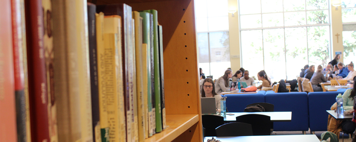 Library books in foreground with students sitting at tables in background.