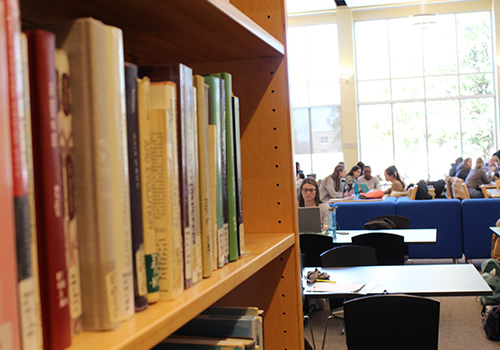 Library books in foreground with students sitting at tables in background.
