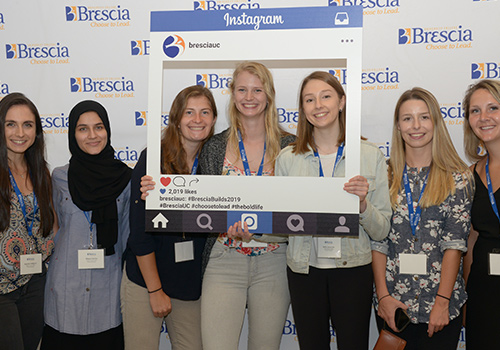 Students standing in front of Brescia selfie wall with Instagram profile cardboard cutout in front of them.
