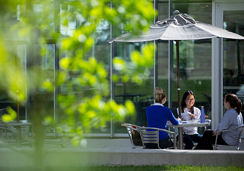 Three students sitting around a table talking and laughing under a patio umbrella on Brescia's campus.